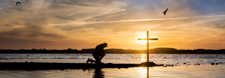 Cross at a lake with a man in prayer beside it, as the sun goes down.